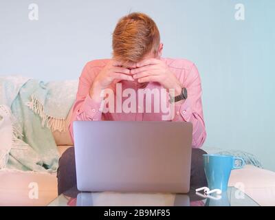 Il biondo uomo d'affari caucasico sta lavorando da casa sul suo computer su un divano con uno sfondo blu chiaro. Foto Stock