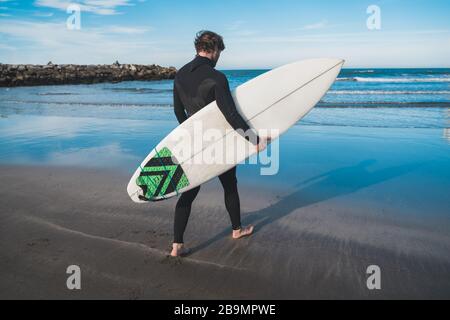 Giovane surfista che entra in acqua con la sua tavola da surf in un costume da surf nero. Concetto di sport e sport acquatici. Foto Stock