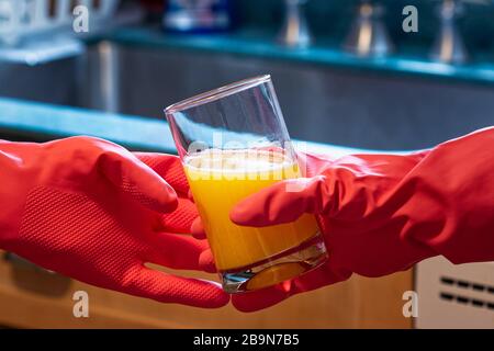 Due persone che indossano guanti in lattice rosso brillante consegnano un bicchiere di succo d'arancia. Sullo sfondo sono elementi di una cucina luminosa. Le immagini illu Foto Stock