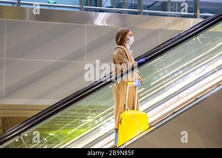 Donna con bagaglio giallo si alza sulla scala mobile, tiene la scala mobile nel terminal dell'aeroporto attraverso guanti in lattice, per proteggersi dal contatto w Foto Stock