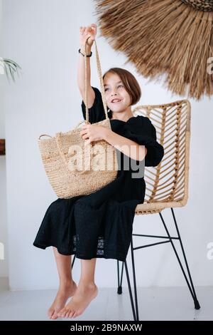 Ragazza curiosa in un abito nero seduta con borsetta in stile tropicale camera Foto Stock