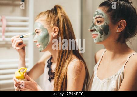 Donna caucasica con i capelli rossi e il suo amico brunette stanno applicando una maschera facciale anti acne mentre guarda nello specchio Foto Stock