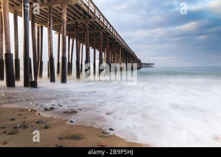 Ventura Pier, Ventura, California. Luce precoce dall'alba visibile da sinistra. Sabbia e rocce in primo piano; onda in arrivo liscia che raggiunge la riva. Foto Stock