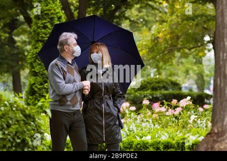 Uomo anziano e donna in maschera facciale. Epidemia di virus. Coppia pensionato che cammina in un parco sotto quarantena durante lo scoppio del coronavirus. Maschere chirurgiche per pr Foto Stock
