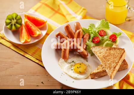 Un piatto per la colazione con salsicce da cocktail, pane tostato, uova fritte, insalate, frutta e un bicchiere di succo d'arancia Foto Stock