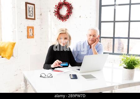 High Angle shot di shopping di coppia senior felice online. Donna anziana attraente che tiene la carta di banca nella sua mano mentre l'uomo anziano che siede dietro la loro l Foto Stock