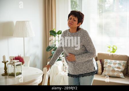 Donna matura che soffre di mal di stomaco a casa. Afferrare e schiacciare il ventre con la mano, sensazione di esausto, in piedi nel soggiorno. Esprimere il dolore Foto Stock