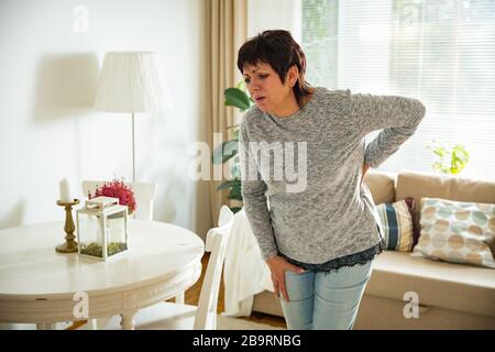 Donna matura che soffrono di mal di schiena a casa. Massaggio schiena inferiore con la mano, sensazione di esaurimento, in piedi nel salotto. Foto Stock