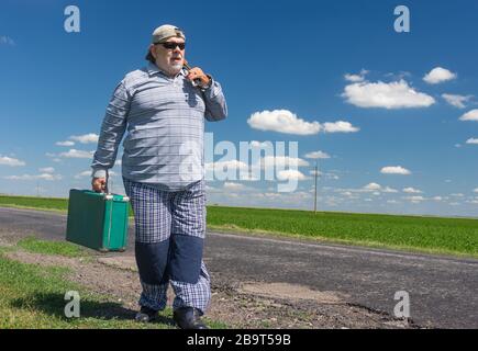 Uomo anziano al bearded che indossa occhiali da sole neri con antica valigia verde nella mano destra e mandolino sulla spalla sinistra che cammina su una strada di campagna Foto Stock