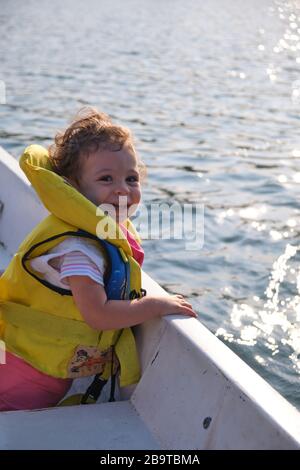 Bambino piccolo guardando la macchina fotografica e sorridendo mentre è su una barca in acqua. Il sole si reflectiing nell'acqua durante il tramonto. Foto Stock