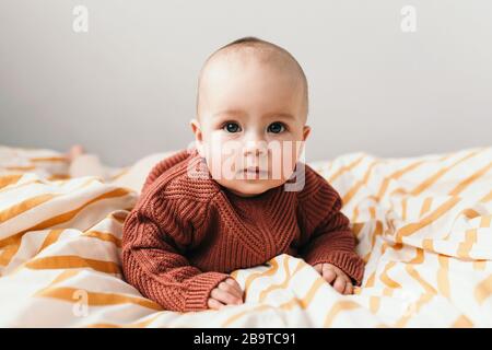 Bella bambina sul letto in un'accogliente maglione marrone sorride. Concetto di maternità e di infanzia. Adorabile bambina di sei mesi sdraiata sul male e guardando nella fotocamera. Foto Stock