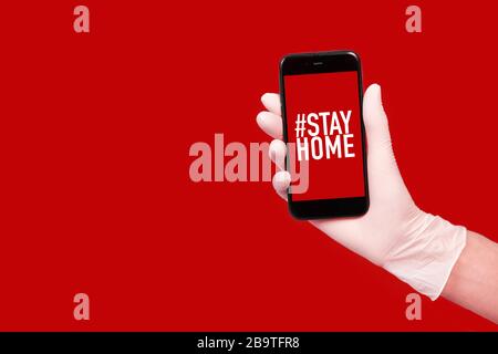 Una mano in un guanto tiene un telefono con le parole rimanere a casa isolata. Quarantena Foto Stock