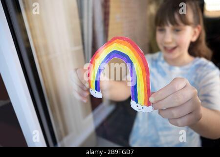 Ragazza che mette l'immagine dell'arcobaleno nella finestra a casa durante la Pandemia di Coronavirus per intrattenere i bambini Foto Stock