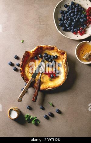 Freschi di forno olandese pancake bambino nella piastra ceramica servita con blackberry e rosse bacche Ribes, ciotola di miele, brocca di crema, vintage posate su beig Foto Stock