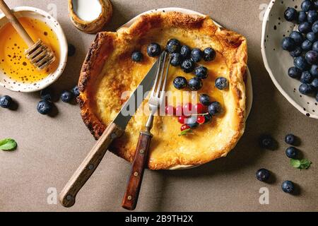 Freschi di forno olandese pancake bambino nella piastra ceramica servita con blackberry e rosse bacche Ribes, ciotola di miele, brocca di crema, vintage posate su beig Foto Stock