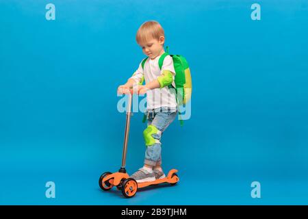 Un ragazzo dai capelli biondi di 4 anni, un bambino con uno zaino e in jeans con una T-shirt bianca guida su uno scooter arancione con protezione verde chiaro su Foto Stock
