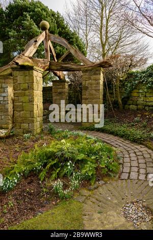 Angolo del giardino cottage paese (giardino rustico, travi in legno, pietre, percorso curvo, miglio, gocce di neve al confine) - York Gate Garden, Leeds UK Foto Stock