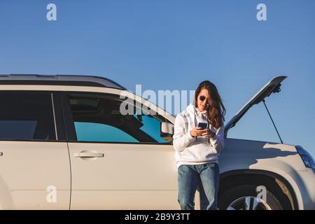 donna in piedi vicino all'auto con cofano aperto al tramonto che parla al telefono Foto Stock