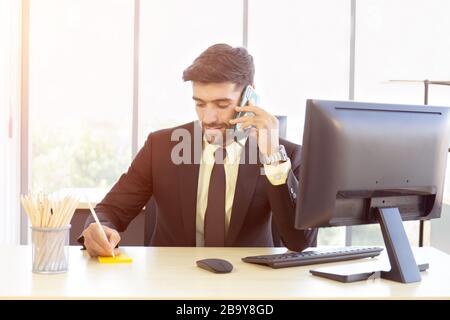 Un uomo d'affari con un abito vestito ordinatamente seduto al telefono con un sorriso luminoso in ufficio Foto Stock