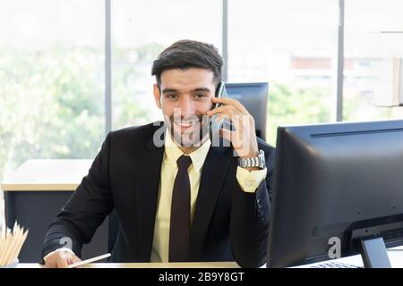 Un uomo d'affari con un abito vestito ordinatamente seduto al telefono con un sorriso luminoso in ufficio Foto Stock
