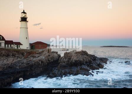 Il faro di Portland Head guida la nave da pesca fuori dalla baia di casco mentre il sole tramonta nel Maine. La torre splende sulla costa rocciosa. Foto Stock