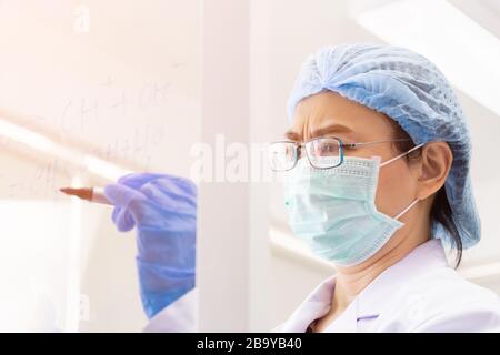 Una scienziata asiatica femminile sta scrivendo la formula per calcolare la chimica su vetro trasparente in un laboratorio. Foto Stock