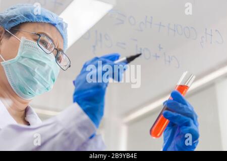 Una scienziata asiatica femminile sta scrivendo la formula per calcolare la chimica su vetro trasparente in un laboratorio. Foto Stock