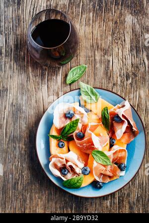 Prosciutto a fette con melone, foglie di basilico e mirtilli serviti con un bicchiere di vino rosso, vista dall'alto Foto Stock