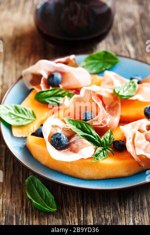 Prosciutto a fette con melone, foglie di basilico e mirtilli serviti con un bicchiere di vino rosso Foto Stock