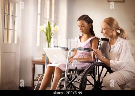 Giovane ragazza in una sedia a rotelle, mostrando qualcosa di sua nonna su un laptop. Foto Stock