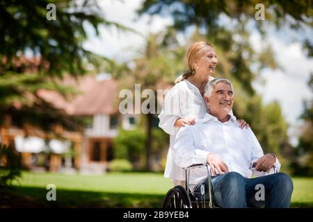 Senior uomo essendo spinto in sedia a rotelle dalla moglie. Foto Stock