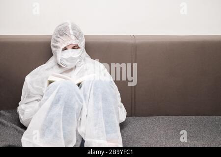 Ragazza in tuta trasparente bianca protettiva, guanti di gomma blu, una maschera medica si siede sul divano a casa e legge il libro. Rimani a casa durante il coronavirus pa Foto Stock