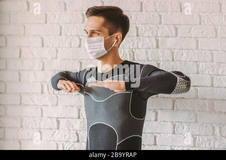 Allenatore professionale di fitness giovane uomo in maschera facciale protettiva facendo esercizi di allenamento. Uomo sportivo in maschera medica sul viso facendo esercizi di stretching Foto Stock