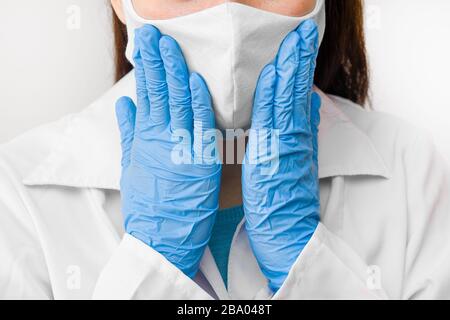 Closeup donne mani al viso in maschera protettiva contro coronavirus, SARS-cov-2, 2019-nCoV o influenza. Medico in camice bianco, guanti blu e maschera. Foto Stock