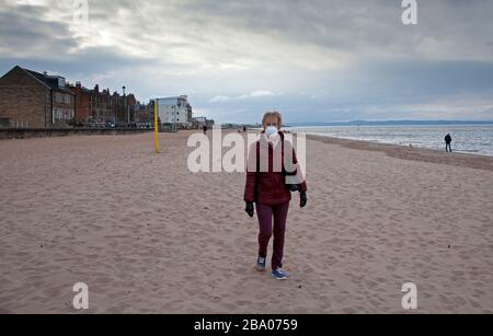 Portobello, Edimburgo, Scozia, Regno Unito. 25th marzo 2020. Pomeriggio estremamente tranquillo sulla spiaggia di Portobello e sul lungomare. Donna anziana nella foto che cammina sulla spiaggia di sabbia indossando una maschera protettiva per la protezione contro il Coronavirus. In Scozia, 719 persone hanno sperimentato un esito positivo per il coroanvirus, con 22 morti. La nuova cifra è un aumento di 135 casi e sei morti durante la notte. Foto Stock