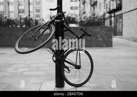 Foto in bianco e nero di una bicicletta incatenata a un lampione a Londra. La bicicletta da corsa è inclinata per montare la serratura di sicurezza. Foto Stock