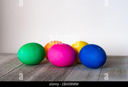 Fila di 5 uova tinte che sono verdi, arancioni, rosa, gialle e blu situate su legno grigio stagionato su uno sfondo bianco Foto Stock