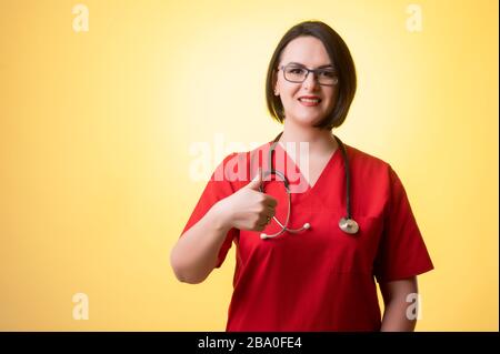 Ritratto di bella donna medico con stetoscopio con scrub rossi, mostrando pollici in posa su uno sfondo isolato giallo. Foto Stock