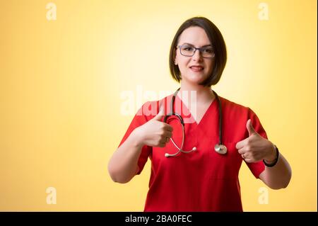 Ritratto di bella donna medico con stetoscopio che porta sctube rosse, mostrando pollici su con entrambe le mani in posa su uno sfondo isolato giallo. Foto Stock