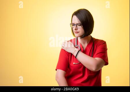 Ritratto di bella donna medico con stetoscopio che porta scrub rossi, ha dolore alle spalle in posa su uno sfondo isolato giallo. Foto Stock