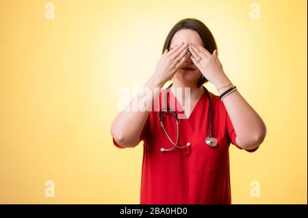 Ritratto di bella donna dottore con stetoscopio con scrub rossi, coprendo occhio come cieco concetto posando su uno sfondo isolato giallo. Foto Stock