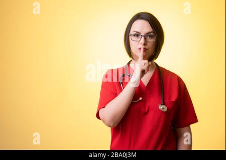 Ritratto di bella donna medico con stetoscopio con scrub rossi, mostrando gesto shh in posa su uno sfondo isolato giallo. Foto Stock