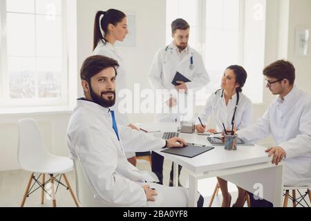 Ritratto di un medico praticante sullo sfondo dei collegi nell'ufficio della clinica. Un medico sicuro è seduto al tavolo in ospedale. Foto Stock