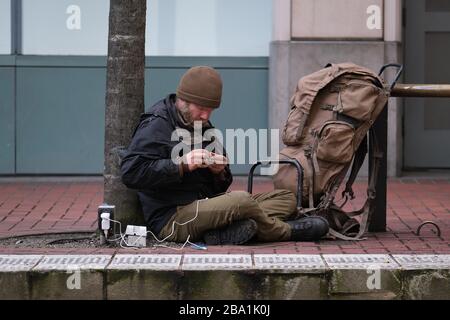 Portland, Stati Uniti. 25 Marzo 2020. Un uomo senza tetto carica il suo telefono fuori Pioneer Place shopping Mall a Portland, Ore., il 25 marzo 2020. I centri commerciali sono uno dei molti affari costretti a chiudere sotto l'ordine esecutivo del Governatore Brown che chiede alle persone di rimanere a casa tranne per i bisogni essenziali, come più estreme misure di distanza sociale mirano a rallentare la diffusione del romanzo coronavirus (COVID-19) e appiattire la curva. (Foto di Alex Milan Tracy/Sipa USA) Credit: Sipa USA/Alamy Live News Foto Stock