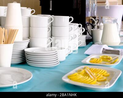 Pile di tazze bianche in ceramica e piattini in mostra. Sulle piastre sono sottili limoni affettati. In una caraffa di vetro di latte. Preparazione per tè e caffè Foto Stock