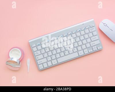 Concetto di blog di bellezza. Uno spazio di lavoro donna, tastiera di un computer e mouse vicino a un contenitore viso crema isolato su uno sfondo rosa pesca con copia spa Foto Stock