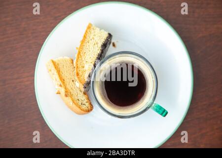 Caffè espresso con crema in tazza di vetro verde su un piattino bianco e verde posto accanto a 2 biscotti con fondo in legno scuro Foto Stock