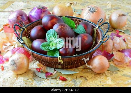 Uova di Pasqua in cesta di vimini e cipolla gialla e rossa, peperoni e basilico verde e foglie di balsamo al limone su sfondo di legno, primo piano Foto Stock