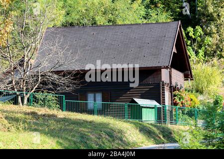 Vista laterale di recente costruzione piccola cabina di legno di montagna marrone scuro circondata da recinzione di metallo verde e fitta vegetazione forestale Foto Stock