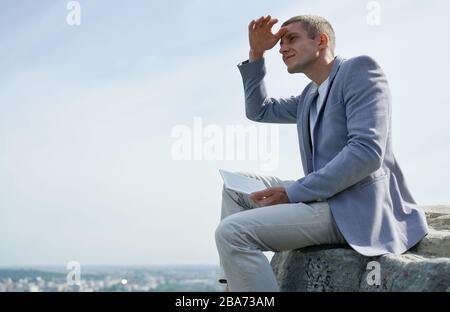 uomo d'affari con un tablet in mano siede su una roccia sullo sfondo della città e guarda in lontananza Foto Stock